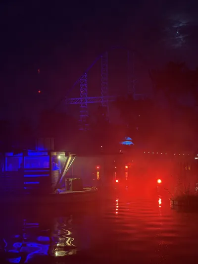 Red and blue light cutting through heavy fog over the water in Cedar Point's Crystal Rock Massacre scare zone, with a coaster hill silhouetted behind the trees