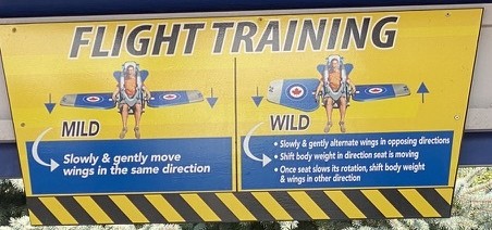 Skyhawk's Flight Training sign at the ride entrance. The MILD column reads: slowly and gently move wings in the same direction. The WILD column reads: slowly and gently alternate wings in opposing directions; shift body weight in the direction the seat is moving; once the seat slows its rotation, shift body weight and wings in the other direction.